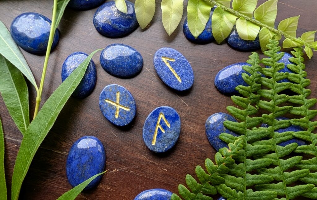 Hand-carved Blue Lapis Lazuli Elder Futhark rune set with the Ansuz, Gebo, and Algiz runes turned face up on a wooden surface.